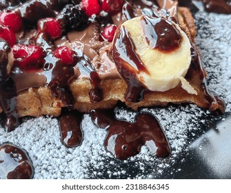 Waffle, dessert. With wild fruit cherry and banana slice. Choco dressing at top and powdered sugar. Black plate. - Powered by Shutterstock
