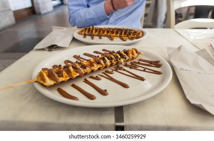 Waffle on a stick with chocolate sauce (coulis) , Milan, Italy. Delicious sweet food served in a ceramic plate (restaurant). Pov with a blurred background. Daylight. July 2019.