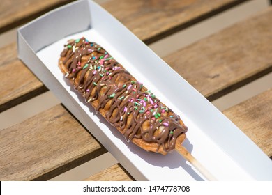 A waffle on a stick, covered with chocolate and colorful candies, served in a paper box