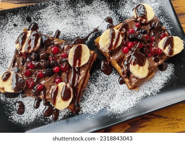 Waffles, with wild fruit, cherry. Choco dressing and banana slices. Powdered sugar. Black plate. Wooden table. - Powered by Shutterstock