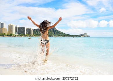 Waikiki beach fun - happy woman on Hawaii vacation. Unrecognizable young adult from behind jumping of joy in water waves, arms up with diamond head mountain in the background, landmark of Honolulu.