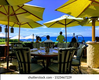 Wailea, Hawaii  United States - November 20 2017 : A Couple Sitting At An Ocean Front Restaurant With A Waiter Standing Beside Them And Many Yellow Patio Umbrellas Overhead.