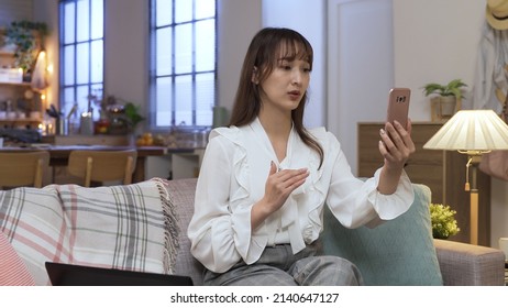 Waist Up Asian Female Ceo Sitting In Living Room With Phone In Hand Making Her Points Clear With Gestures In A Video Conference. Work From Home During Self-quarantine. Technology And Communication