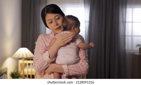 waist up asian mom is embracing and patting on her baby’s back trying to put her to sleep at nighttime in illuminated home interior.