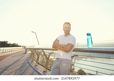 Waist Length Portrait Of An Attractive Middle Aged Muscular Build European Man In Sportswear Standing On City Bridge And Relaxing After Heavy Workout Outdoor. Sunbeams Falling On The Treadmill At Dawn