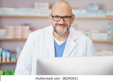 Waist Up Occupation Portrait Of Friendly Mature Male Pharmacist Wearing White Lab Coat And Working On Computer Behind Counter In Drug Store Pharmacy