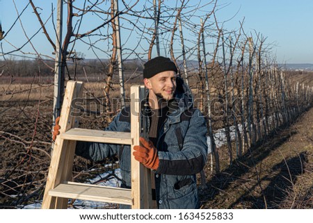 Waist up photo of the young happy man gardener in black hat and orange work gloves that stands among his garden, holds wooden stepladder in his hands and looking at right side, sunny winter day