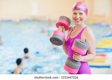 Waist up portrait of active senior woman wearing swimsuit posing by swimming pool holding foam dumbbells and smiling at camera, copy space - Powered by Shutterstock