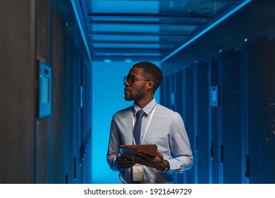 Waist up portrait of African American data engineer holding digital tablet while working with supercomputer in server room lit by blue light, copy space