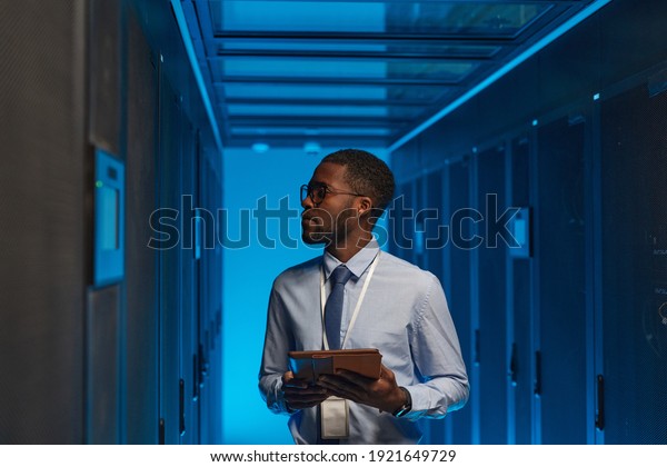 Waist up portrait of African American
data engineer holding digital tablet while working with
supercomputer in server room lit by blue light, copy
space