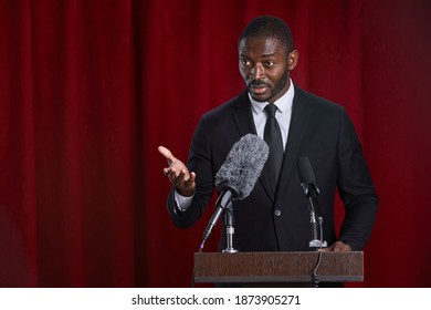 Waist Up Portrait Of African-American Man Speaking To Microphone Standing At Podium On Stage Against Red Curtain, Copy Space