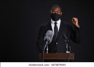 Waist Up Portrait Of African-American Man Wearing Mask While Giving Powerful Speech Standing At Podium Against Black Background, Copy Space