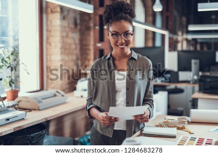 Waist up portrait of attractive young lady in glasses standing near table with notebook, measuring tape and color palette. She is looking at camera and smiling