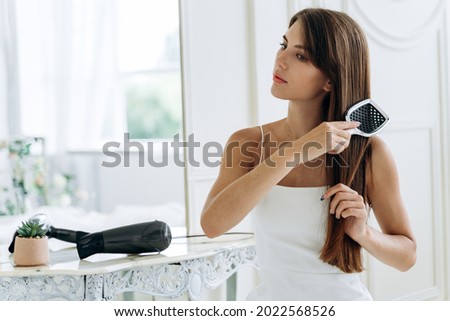 Waist up portrait of beaming lady combing her long dark hair while looking away with calm smile. Hair care concept. Stock photo 