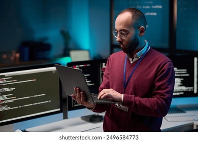 Waist up portrait of bearded adult man holding laptop while standing in cybersecurity office with code lines on screens copy space