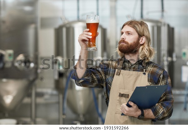 Waist up portrait of bearded brewmaster holding\
beer glass while inspecting quality of production at brewing\
factory, copy space
