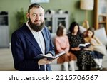 Waist up portrait of bearded jewish man wearing kippah and looking at camera holding book, copy space
