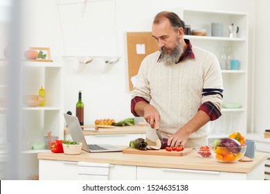 Waist Up Portrait Of Bearded Senior Man Watching Video Recipe Via Laptop While Cooking Healthy Salad In Kitchen, Copy Space