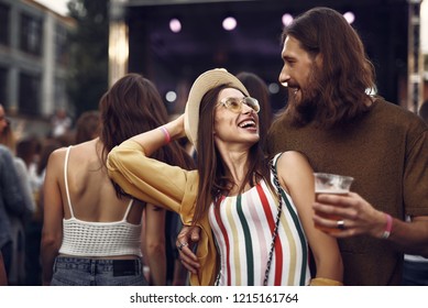 Waist Up Portrait Of Beautiful Couple Enjoying Company Of Each Other At Concert. Girl In Hat Looking At Bearded Man With Beer While He Hugging Her From Behind