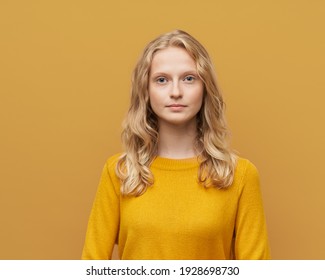 Waist Up Portrait Of Beautiful Young Serious Clever Blonde Scandinavian Woman On Bright Yellow Orange Wall. Fresh Face Of Female Without Makeup Face, Curly Hair, Yellow Jumper, Teen In Orange Sweater