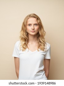 Waist up portrait of beautiful young serious clever blonde woman without makeup on beige wall. Pretty female with curly hair in white t shirt