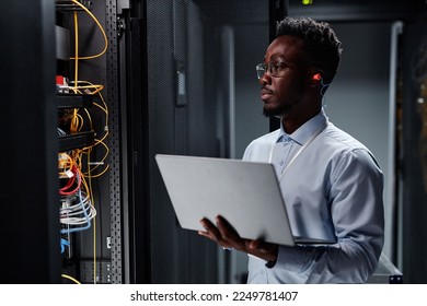 Waist up portrait of black man as network engineer in server room using laptop, minimal