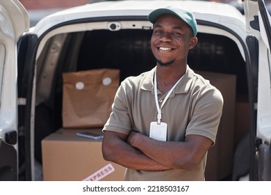 Waist Up Portrait Of Black Young Man As Delivery Man Standing With Arms Crossed By Van In Sunlight And Smiling At Camera