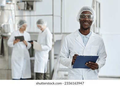 Waist up portrait of black young man wearing lab coat and smiling at camera in clean workshop of pharmaceutical factory, copy space - Powered by Shutterstock