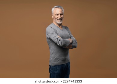 Waist up portrait Caucasian handsome muscular attractive confident gray haired senior man, pensioner smiling a toothy smile, looking at camera, standing with folded arms over isolated brown background - Shutterstock ID 2294741333