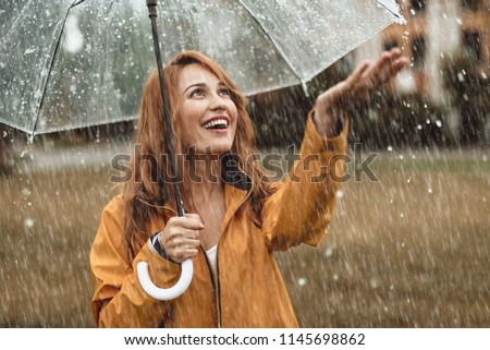 Waist up portrait of cheerful girl holding umbrella and catching rain drops with smile. She is stretching hand and looking upwards with sincere delight