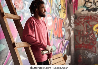 Waist up portrait of confident graffiti writer standing leaning on the ladder and happily smiling while holding a spray paint can and looking away