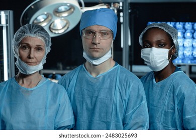 Waist up portrait of diverse surgical team of three people in operating room looking at camera in low light - Powered by Shutterstock