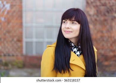 Waist Up Portrait of Fashionable Young Woman with Long Dark Hair Wearing Yellow Coat Outdoors in front of Urban Brick Wall, Looking Up Optimistically to the Side, with Copy Space - Powered by Shutterstock