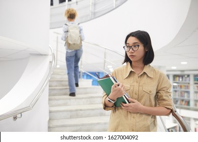 Waist Up Portrait Of Female Asian Student Holding Books While Going Down Spiral Stairs In Modern College Interior, Copy Space