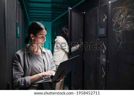 Waist up portrait of female data engineer using laptop in server room while setting up supercomputer network, copy space
