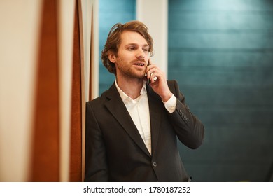 Waist Up Portrait Of Handsome Bearded Businessman Speaking By Smartphone And Looking Away While Leaning On Wall In Minimal Office Interior, Copy Space