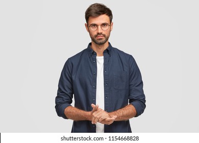 Waist Up Portrait Of Handsome Serious Unshaven Male Keeps Hands Together, Dressed In Dark Blue Shirt, Has Talk With Interlocutor, Stands Against White Background. Self Confident Man Freelancer