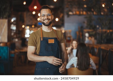 Waist up portrait of handsome waiter smiling cheerfully at camera standing in restaurant or cafe, copy space. Young waiter serving coffee in a cafe and looking at camera. - Powered by Shutterstock