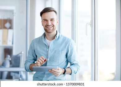 Waist up portrait of handsome young man holding digital tablet and smiling happily at camera standing by window, copy space - Powered by Shutterstock