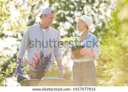 Waist up portrait of happy senior couple walking towards camera in garden pushing cart with plants