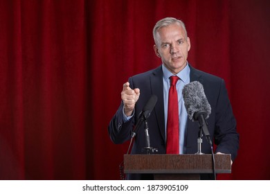 Waist Up Portrait Of Mature Man Speaking To Microphone Standing At Podium On Stage Against Red Curtain, Copy Space