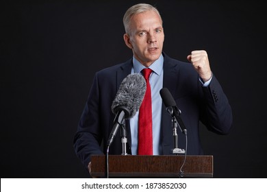 Waist Up Portrait Of Mature Man Standing At Podium And Gesturing While Giving Speech To Microphone Against Black Background, Copy Space