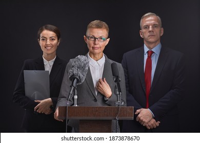 Waist Up Portrait Of Mature Woman Giving Speech Standing At Podium With Two Assistants In Background