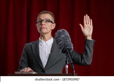 Waist Up Portrait Of Mature Woman Giving Oath While Standing At Podium On Stage Against Red Curtain, Copy Space