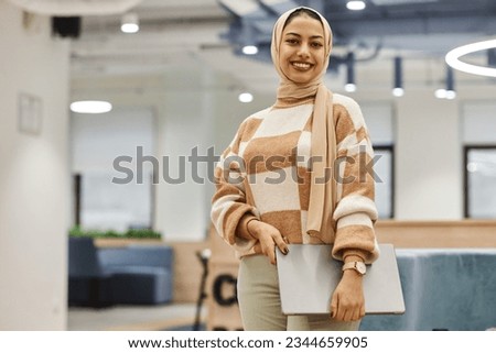 Waist up portrait of modern Middle Eastern woman wearing head cover while standing in office interior and smiling at camera cheerfully, copy space