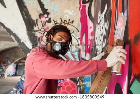 Waist up portrait of multiracial man with dreadlocks wearing respirator painting colored graffiti on public space wall while standing at the stepladder and looking to the camera