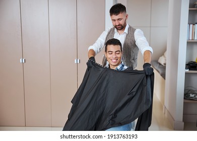 Waist Up Portrait Of Pleasant Indian Man In Shirt Sitting On The Chair While Barber Closing His Client With Black Peignoir In Beauty Salon