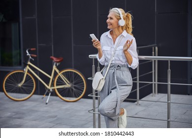 Waist Up Portrait Of Pretty Lady Talking With Friends On Mobile Phone While She Is Standing In Front Of Modern Building And Retro Bike