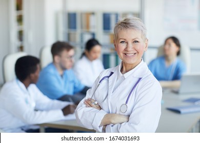 Waist Up Portrait Of Senior Female Doctor Standing With Arms Crossed And Smiling At Camera Against Medical Conference Background, Copy Space