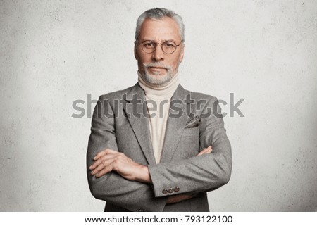 Waist up portrait of serious bearded mature man keeps arms folded, dressed in formal suit, confident in his knowledge, isolated on white concrete background. Company director comes on business meeting